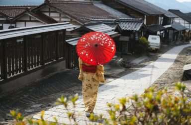Woman in kimono with red umbrella walking through a traditional Japanese village street.